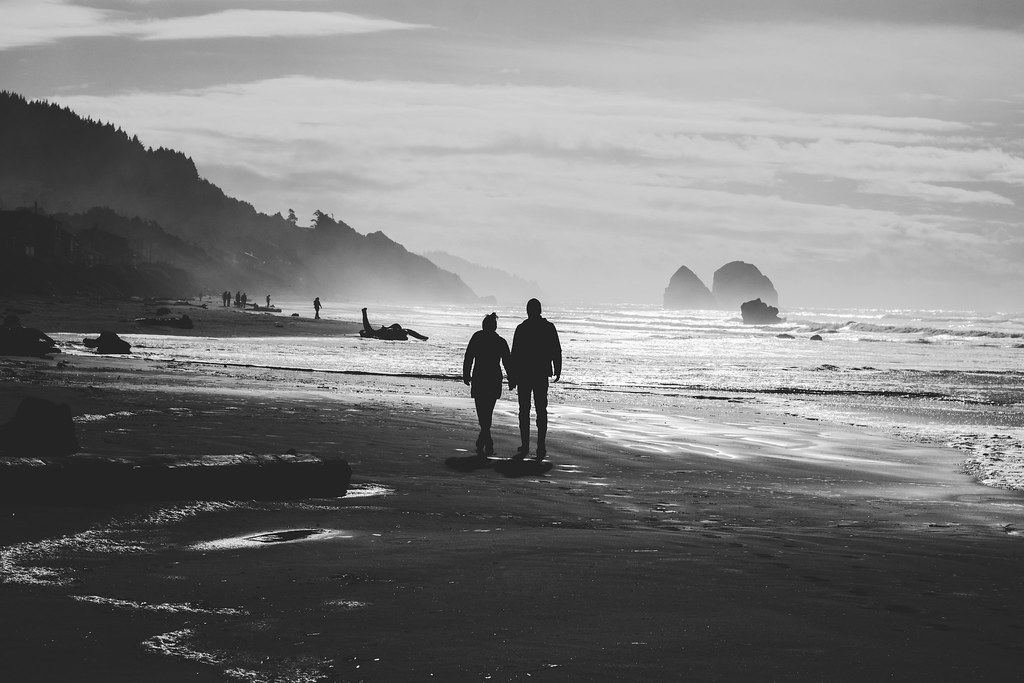 Two people walking along an empty beach, in black and white