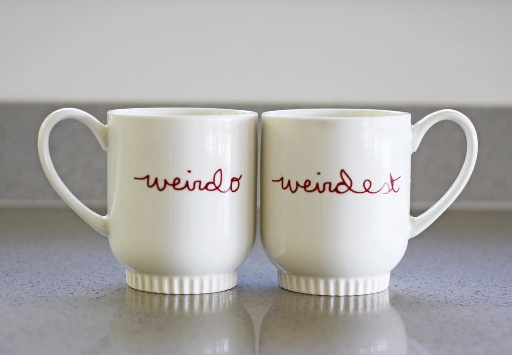 Two white mugs side by side, one says weirdo, the other weirdest
